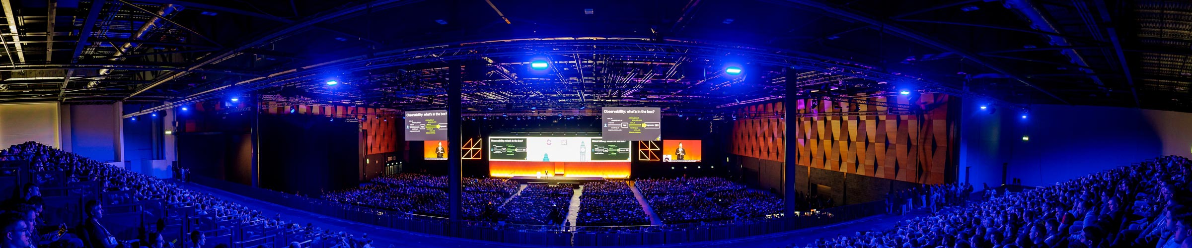 Panoramic view of a packed keynote session in a large, dimly lit conference hall, with colorful lighting and multiple large screens displaying a presentation on observability.